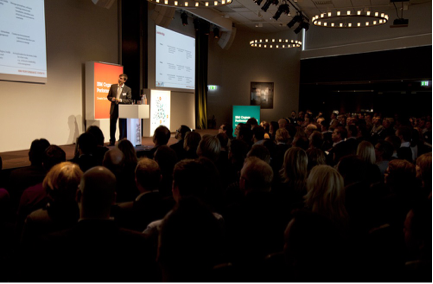 Mika delivering a presentation at a conference podium to a large, attentive audience in a dimly lit auditorium. Stage with orange accents and dual projection screens visible.